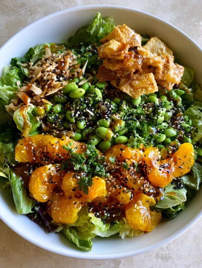 Close-up of a large white ceramic bowl filled with crisp romaine, edamame, golden wonton strips, and mandarin oranges coated in glossy Asian Sesame Salad Dressing.