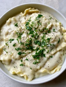 Close up shot of glossy white Garlic Parmesan Ravioli Sauce with visible black pepper flecks and fresh parsley on square ravioli.