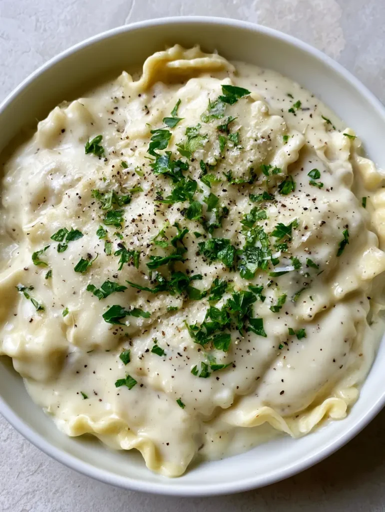 Close up shot of glossy white Garlic Parmesan Ravioli Sauce with visible black pepper flecks and fresh parsley on square ravioli.