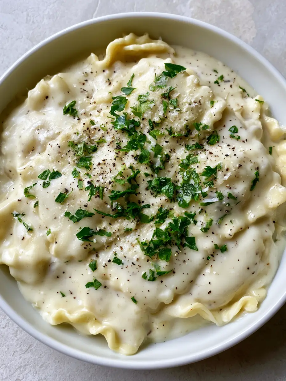 Close up shot of glossy white Garlic Parmesan Ravioli Sauce with visible black pepper flecks and fresh parsley on square ravioli.