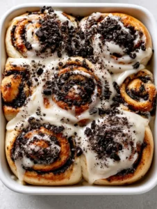 Close-up of golden-brown yeast dough rolls with dark chocolate cookie filling and glossy white cream cheese glaze. (Cookies and Cream Cinnamon Rolls)