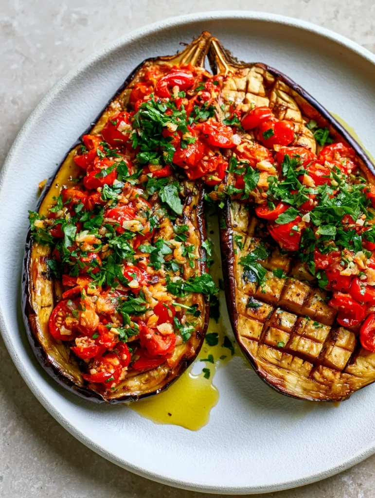 A pair of roasted eggplant halves on a white ceramic plate topped with roasted garlic and blistered red tomatoes.