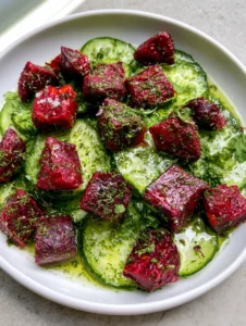 A close-up top-down shot of a shallow white ceramic bowl filled with thinly sliced cucumbers and matte roasted beetroot cubes. (Cucumber Beetroot Salad)