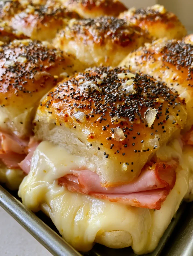 A metal tray filled with 12 Hawaiian Ham and Cheese Sliders speckled with poppy seeds and onions.