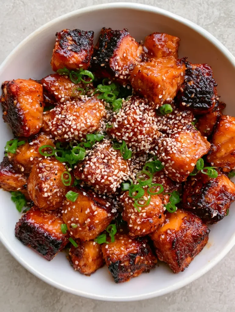 Top-down view of a white ceramic bowl with salmon bites garnished with green scallions and sesame seeds.