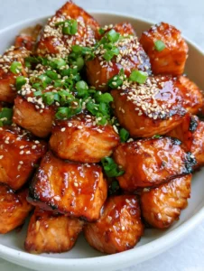 A handheld smartphone photo of crispy honey garlic salmon bites piled in a shallow white ceramic bowl with sesame seeds and scallions. (Honey Garlic Salmon Bites)